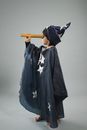 Full body side view of barefoot boy in astrologer outfit with hat looking through telescope on gray background in studio