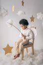 Full body of barefoot African American boy wearing white wings sitting on chair with decorative yellow star on wand in hands on white background in studio with cotton wool