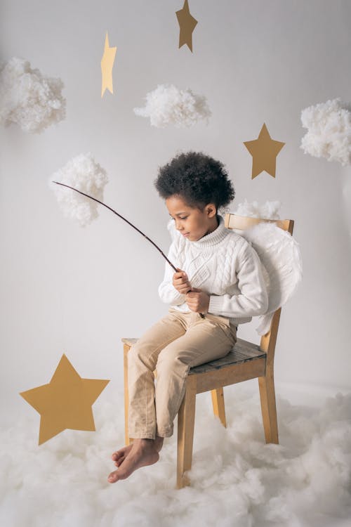 Full body of barefoot African American boy wearing white wings sitting on chair with decorative yellow star on wand in hands on white background in studio with cotton wool