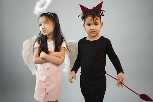 Adorable Asian girl in angel costume with halo standing near black boy in devil outfit looking at camera on gray background in studio