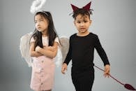 Adorable Asian girl in angel costume with halo standing near black boy in devil outfit looking at camera on gray background in studio