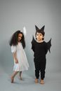 African American girl in white dress and angel halo headdress near barefoot boy in bat costume on gray background