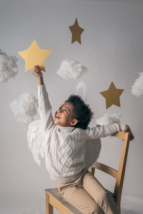 Cheerful African American child with angel wings touching decorative star while looking up from chair on light background