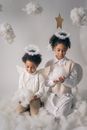 Ethnic sister and brother in angel costumes touching decorative clouds
