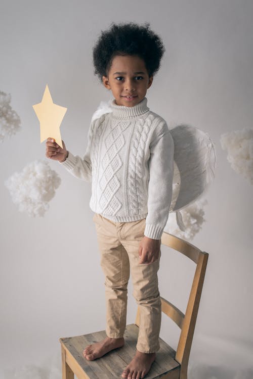 Full body of African American boy in warm sweater and angel wings standing on chair with paper star against fluffy clouds made of cotton wool
