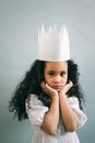 Sweet little African American girl with curly hair wearing white dress and paper crown for Halloween celebration against gray background