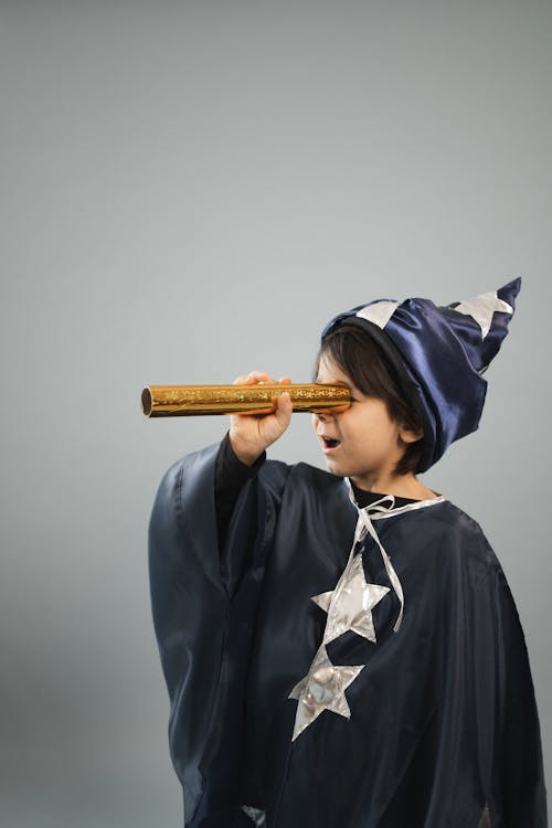 Surprised boy wearing masquerade costume of magician looking through kaleidoscope tube while standing against gray background