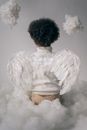 Back view of faceless boy in white angel wings sitting on cotton wool on white background in light studio with decorated hanging clouds