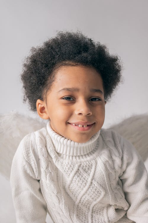 Happy African American boy in warm sweater wearing angel wings smiling and looking at camera on white background in studio