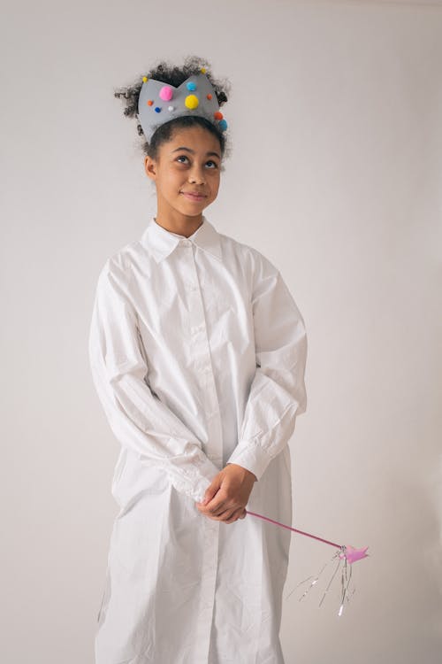 Dreamy African American female wearing crown with pompoms looking up while standing on white background with magic wand in hand