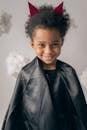 Cheerful African American boy wearing black devil with red horns looking at camera while standing on white background with decorative cotton clouds