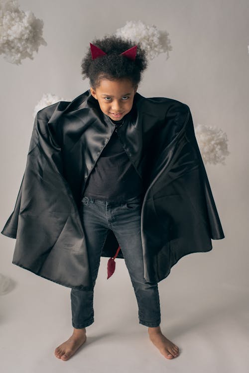 Full body of little African American girl with afro in black leather outfit and red devils horns standing on white background