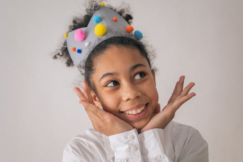 Happy African American girl with curly hair and gray crown with colorful pompoms smiling and looking away with hands at chin on white background