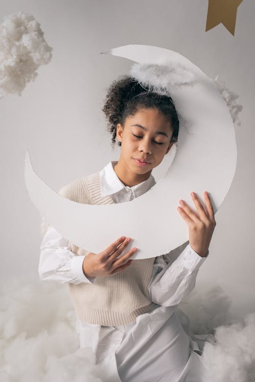 Cute little girl in white clothes and nimbus holding paper moon with eyes closed while sitting on cotton on white background in studio