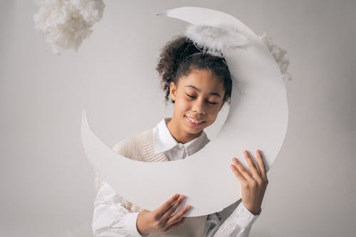 Smiling African American girl in white outfit and nimbus holding white paper moon on white background with cotton in studio