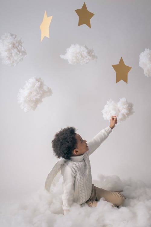 Free Little boy in trendy warm clothes and angel wings sitting on floor and touching handmade clouds from cotton on white background in studio Stock Photo