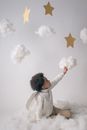 Little boy in trendy warm clothes and angel wings sitting on floor and touching handmade clouds from cotton on white background in studio