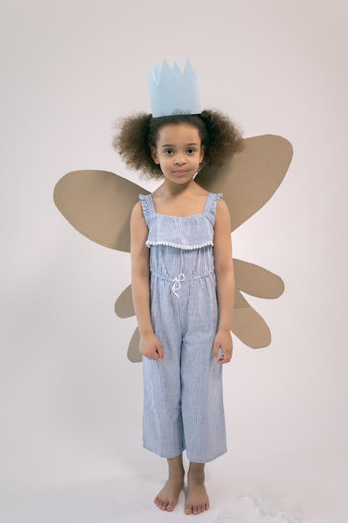 Full body of adorable little girl with paper crown and cardboard wings looking at camera and standing on white background in studio