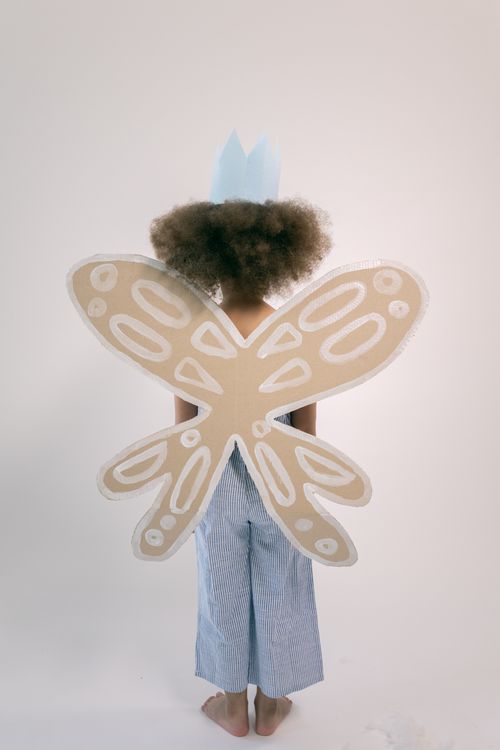 Free Back view full body of little girl with afro wearing blue paper crown and cardboard wings with white pattern while standing in white studio Stock Photo