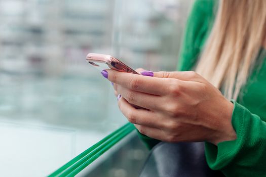 Crop unrecognizable blond female with manicure text messaging on cellphone against window on blurred background