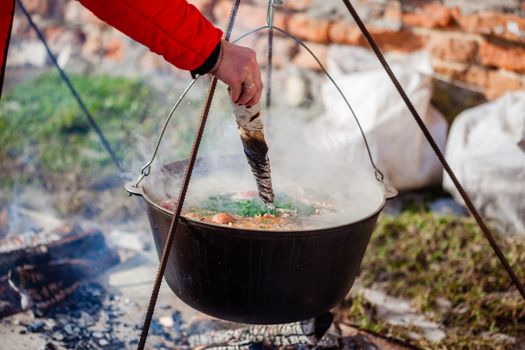 Crop unrecognizable person immersing birch firewood into ukha in cauldron with smoke in daylight on blurred background