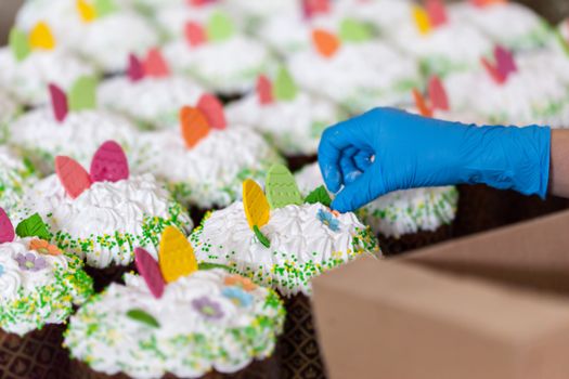 Crop unrecognizable worker in disposable glove with box against tasty desserts with egg and floral decor on whipped meringue cream