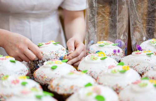 Crop anonymous female confectioner preparing Easter cakes decorated with colorful sprinkles and sweet glaze