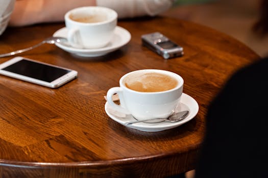 High angle of mugs with latte on saucer with spoon placed on wooden table near phone and voice recorder near crop anonymous person in light cafeteria