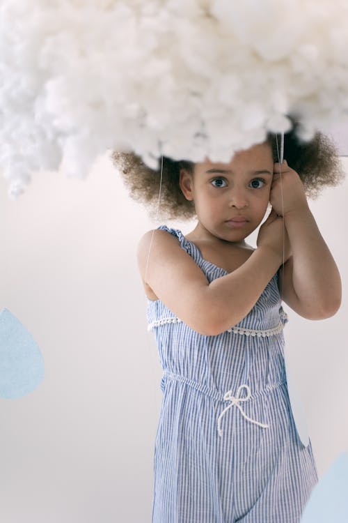 Adorable little African American girl with curly hair in blue dress standing under handmade cloud from cotton in white studio