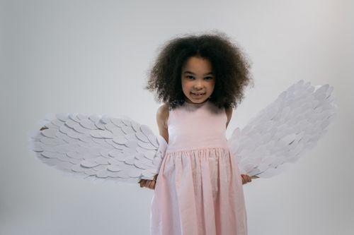 Free Adorable little ethnic girl with dark Afro hair in stylish pink dress and angel wings smiling and looking at camera against white background Stock Photo