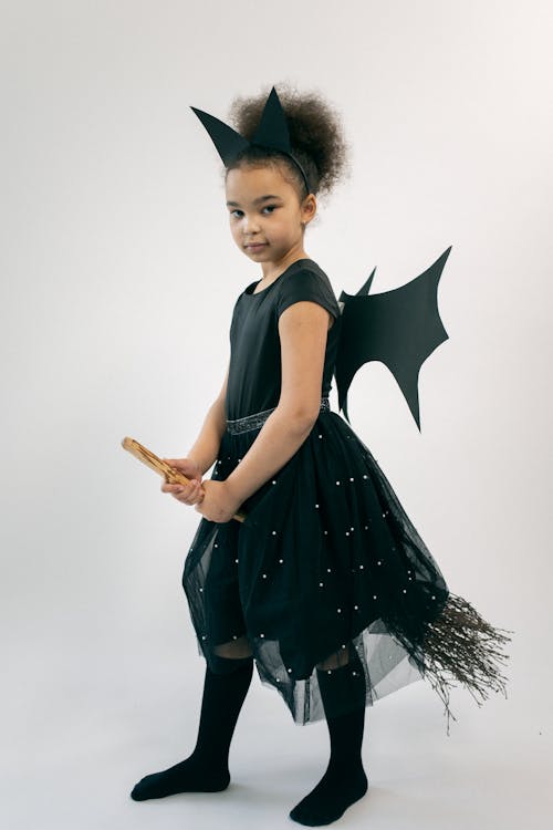 Full body of adorable ethnic little girl in witch costume and wings holding broom and looking at camera against white background