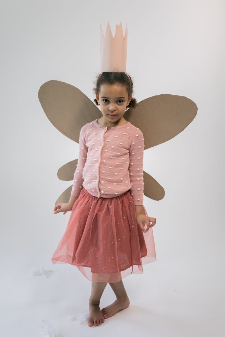 Cute Black Kid With Paper Crown And Butterfly Wings Standing In White Studio