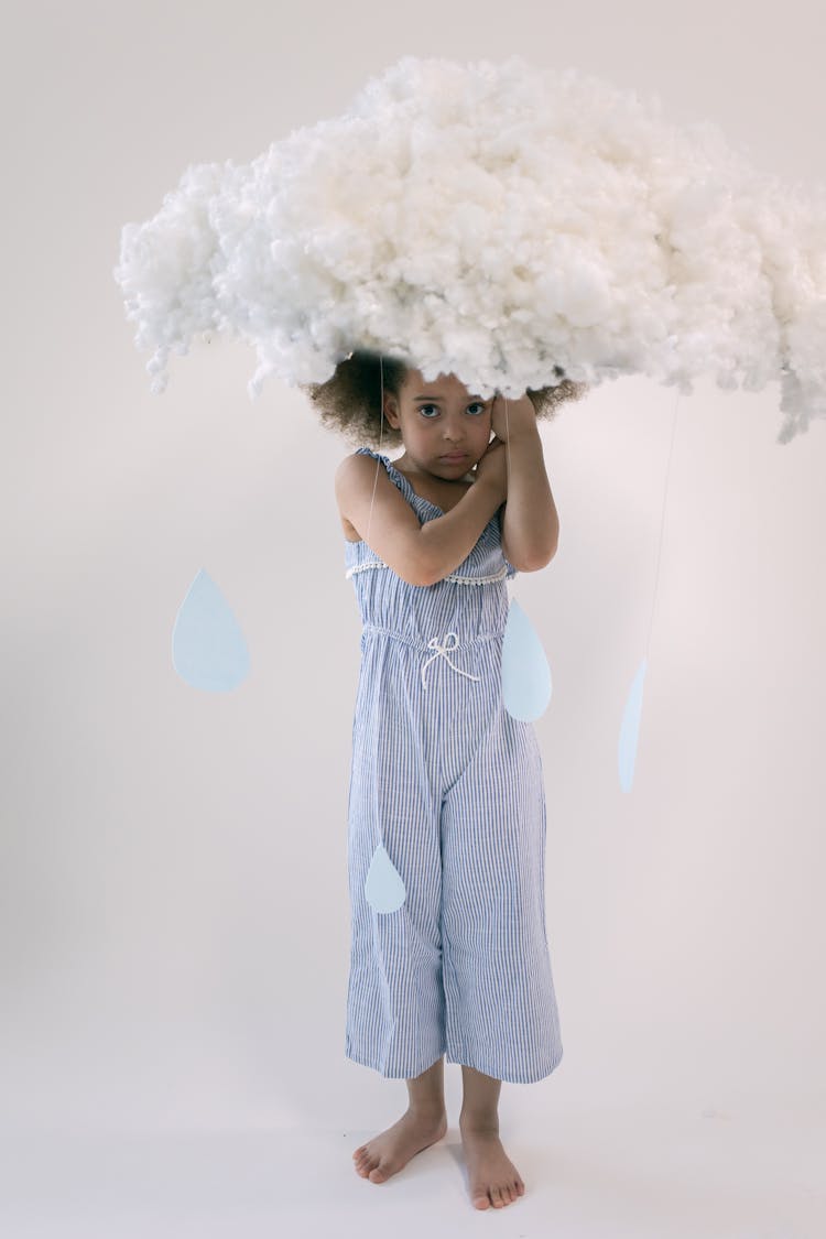 Upset Little Ethnic Girl Standing Under Artificial Cloud In White Studio