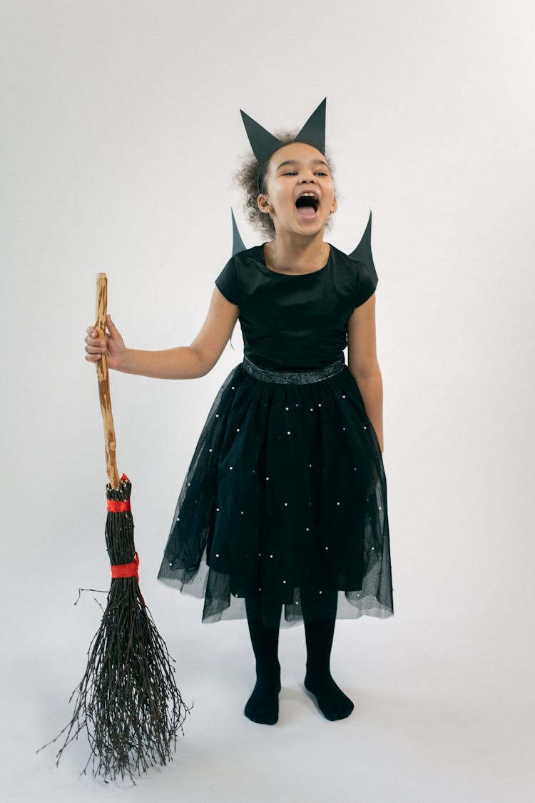 Cute Little Black Girl In Halloween Costume Standing In Studio With Broom And Screaming