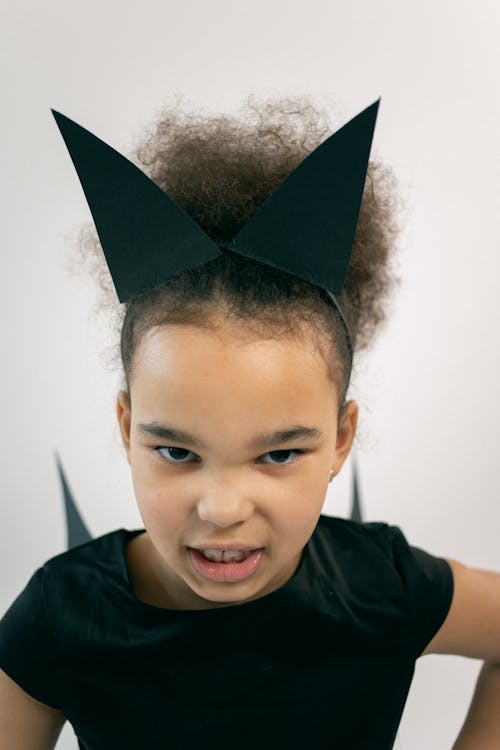 Free Serious little black girl with Afro hair in Halloween costume and headband gazing at camera and roaring while imitating cat against white background Stock Photo
