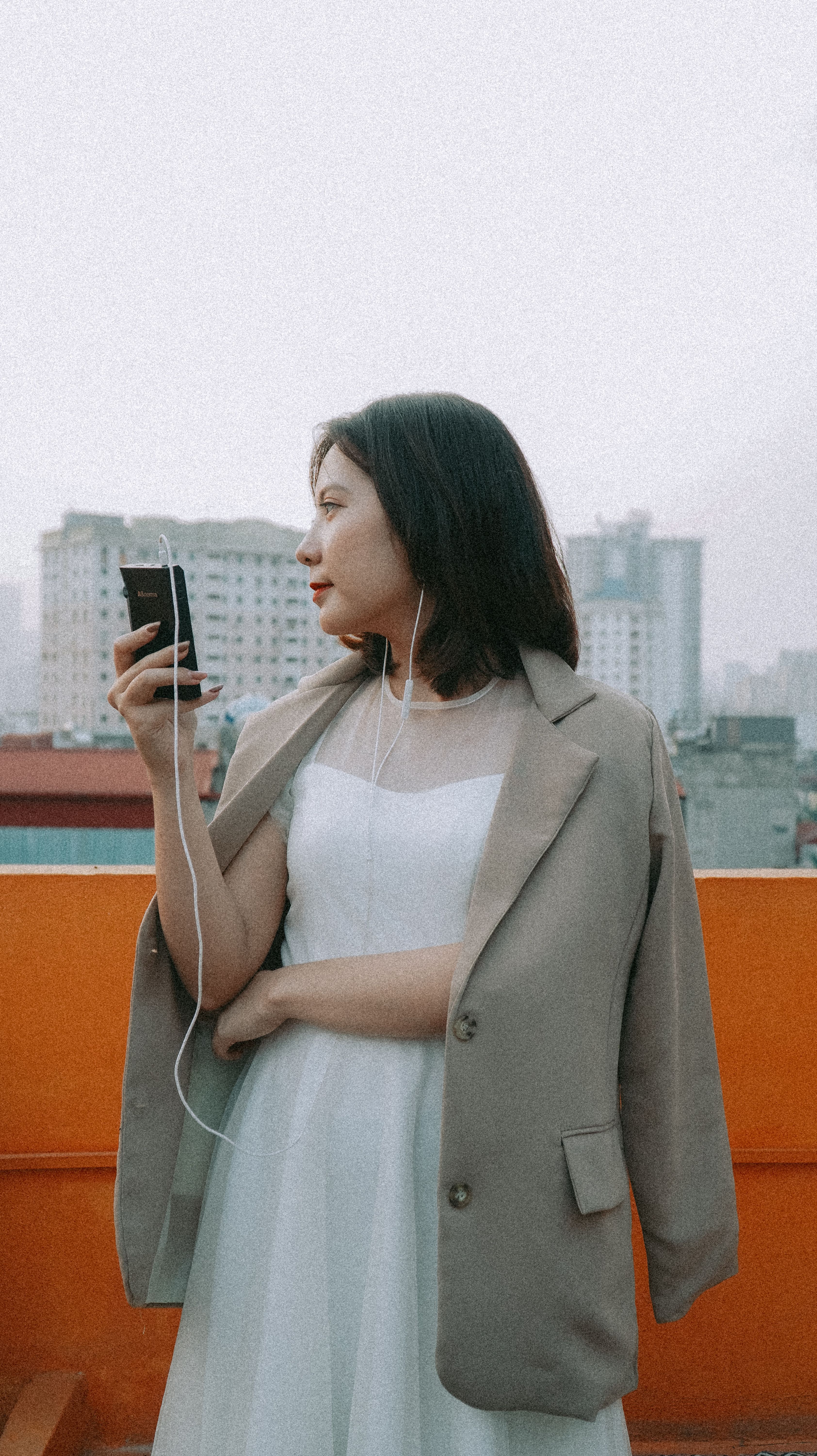 A young woman in a white dress and blazer enjoys music with earphones on a rooftop.