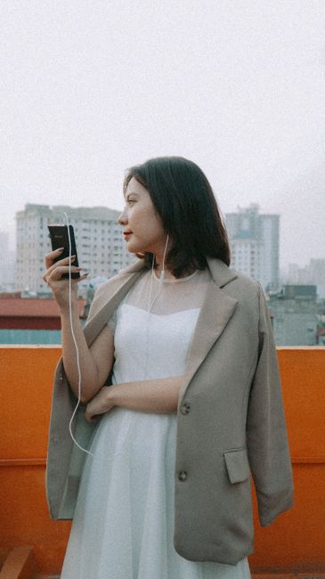A young woman in a white dress and blazer enjoys music with earphones on a rooftop.