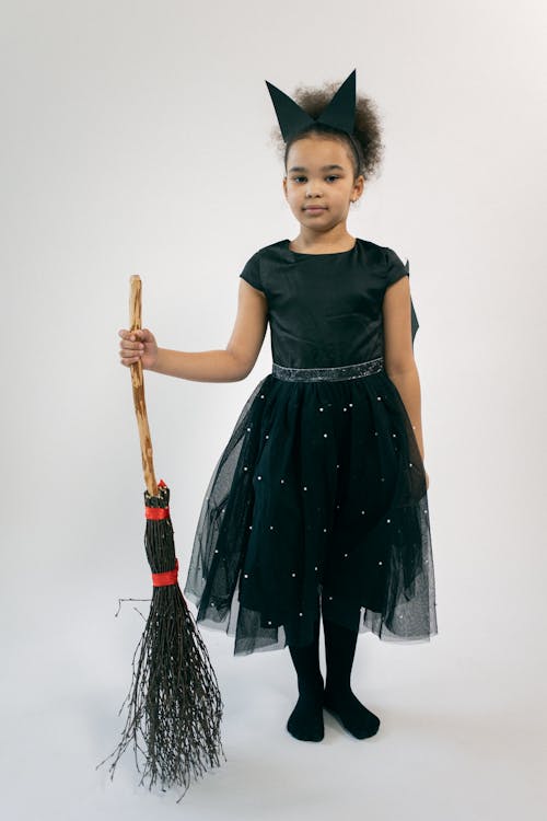 Cute black kid in witch costume with broom in hand looking at camera