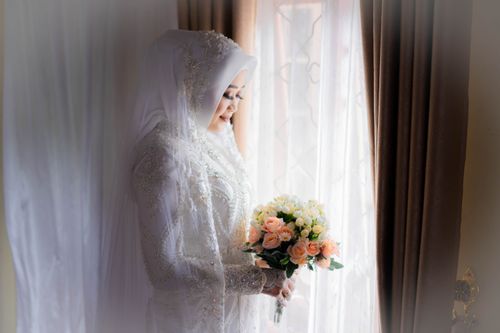 Free A beautiful bride in a white dress holding a bouquet by a window, captured with soft natural light. Stock Photo