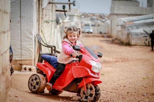Free A smiling girl rides her red toy motorcycle on a dirt road with buildings around her in Syria. Stock Photo