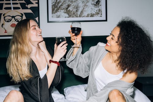 Free Two Young Women Sitting on White Bed While Drinking Their Red Wine Stock Photo