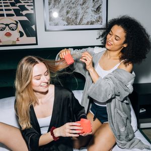 Two laughing friends on a bed at a sleepover, one putting coral rollers into the other's long hair