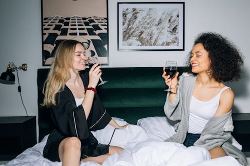 Free Two Young Women Sitting on White Bed While Drinking Their Red Wine Stock Photo
