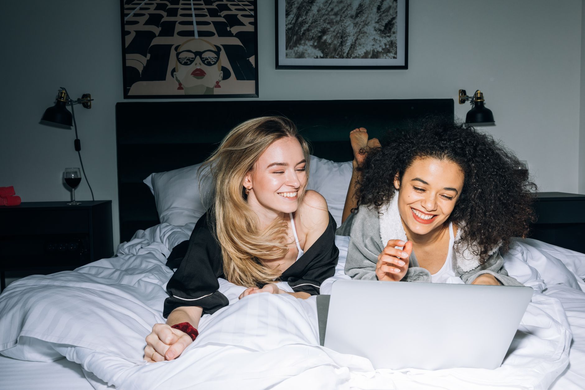 청년지원금 해지 시 불이익 3가지 주요 사례 6 Two young women smiling and lying on a bed, enjoying a laptop together in a cozy bedroom setting.
