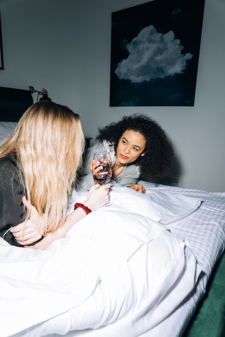 Two Young Women Lying On White Bed While Looking At Each Other