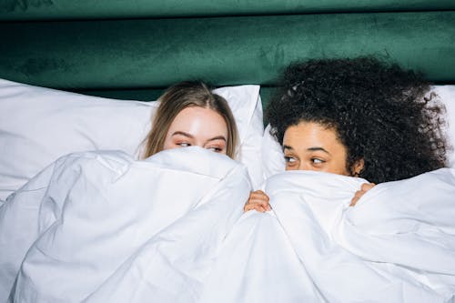 Free Two Young Women Lying on White Bed While Looking at Each Other Stock Photo