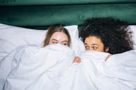 Two Young Women Lying on White Bed While Looking at Each Other