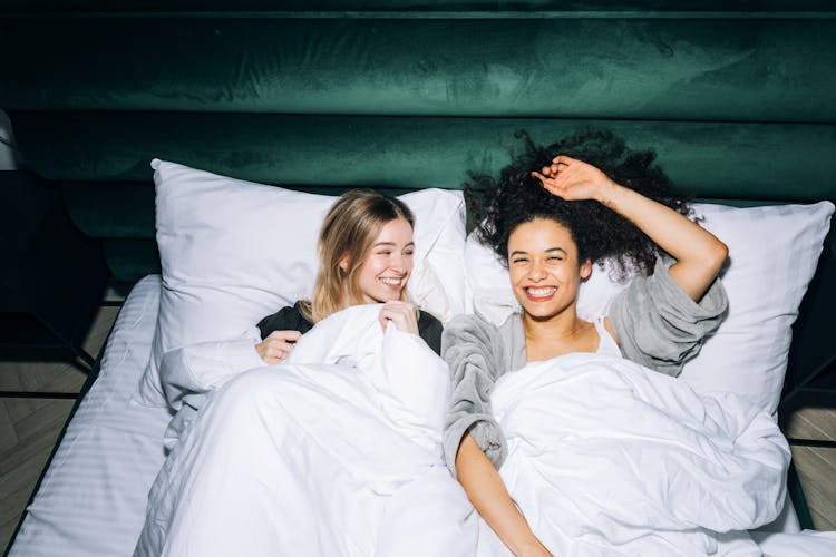 Two Young Women Having Fun While Lying On White Bed