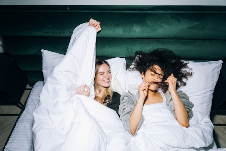 Two Young Women Having Fun While Lying On White Bed