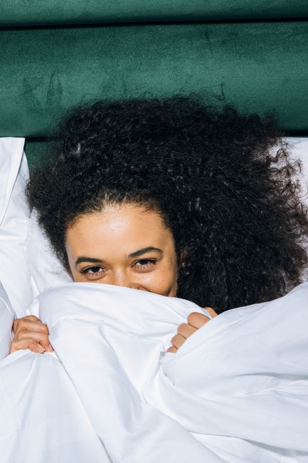 A joyful young woman with afro hair peeks from under a white comforter in a cozy bedroom.
